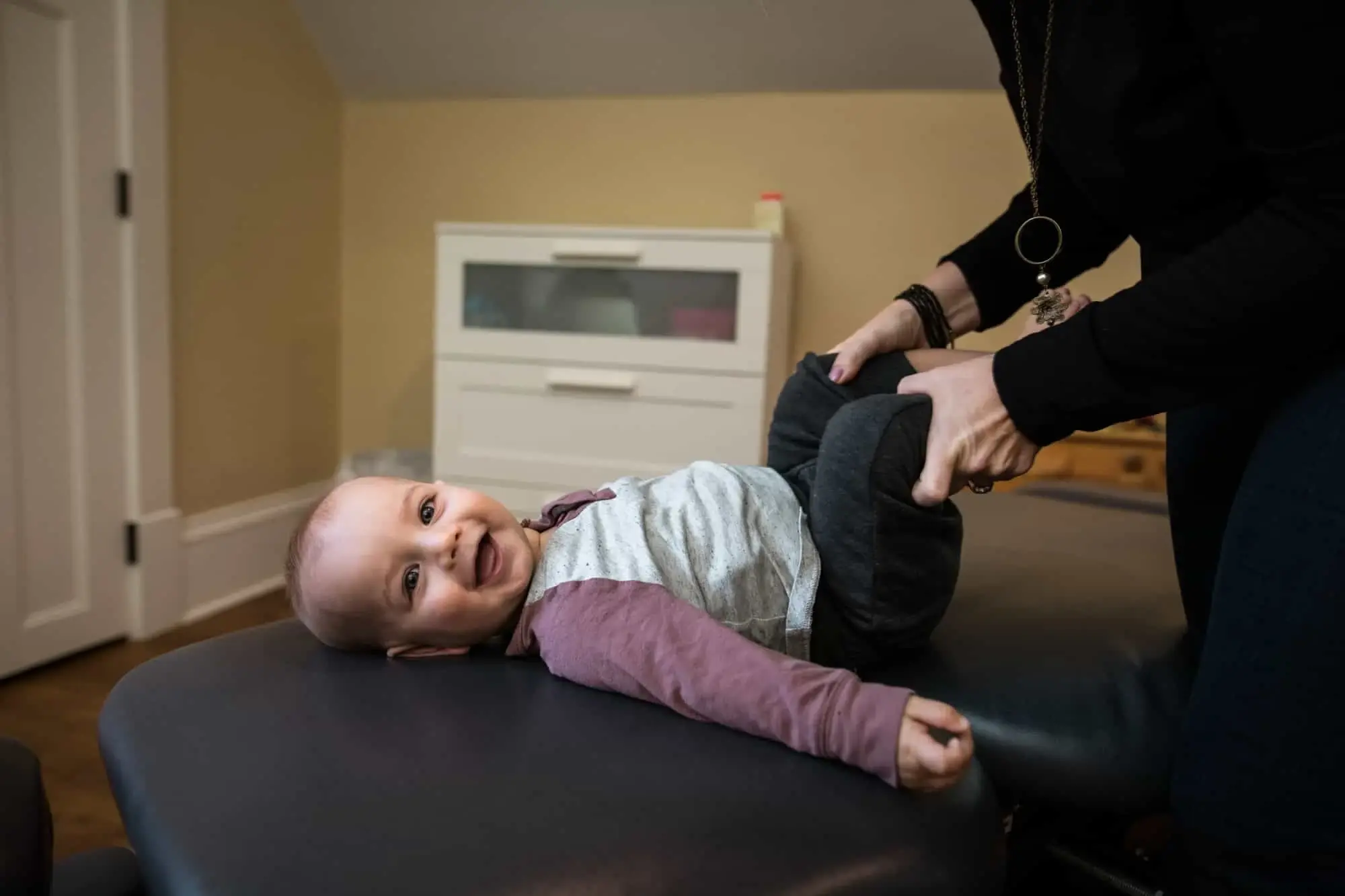 A smiling baby lies on an adjusting table while a doctor gently supports their legs